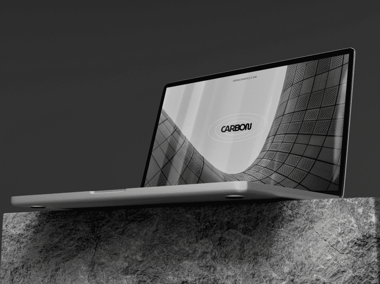 Project-Pic-1png - Corporate video production company in Delhi Slim silver laptop resting on a rough rock surface, screen shows a grayscale curved architectural image with a CARBON logo.
